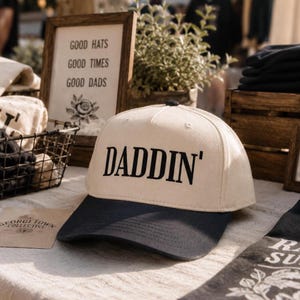 May include: A two-tone baseball cap featuring a cream-colored crown and a dark navy blue brim. The front of the crown displays the word DADDIN' printed in bold, black, capital letters. The cap has a structured five-panel design with a top button.