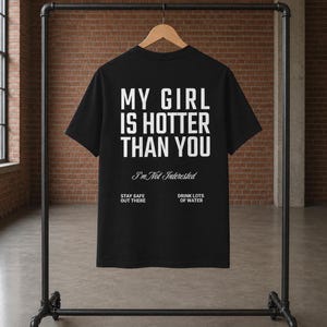 May include: A black short-sleeved t-shirt featuring white text that reads MY GIRL IS HOTTER THAN YOU, followed by I'm Not Interested in script, and at the bottom, STAY SAFE OUT THERE and DRINK LOTS OF WATER in a clean, sans-serif font.