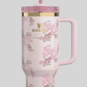 Puede incluir: Un vaso Stanley de color rosa claro con un estampado de flores y lazos en tonos rosa y verde. El vaso incluye un borde dorado, un asa rosa a juego, una tapa transl&uacute;cida y una pajita rematada con un accesorio de lazo rosa decorativo.