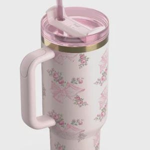 Puede incluir: Un vaso t&eacute;rmico de color rosa claro con asa, que presenta un patr&oacute;n repetitivo de lazos rosas y peque&ntilde;os grupos de flores. El vaso incluye una tapa transparente con una pajita rosa a juego rematada con un lazo decorativo y un borde dorado.