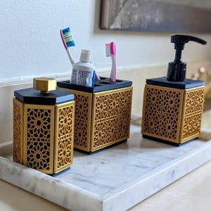 Puede incluir: Un juego de accesorios de baño sobre una bandeja rectangular de mármol blanco, que incluye un recipiente con tapa, un soporte para cepillos de dientes con dos cepillos y un dispensador de jabón con bomba negra. Cada pieza tiene base negra y paneles dorados.