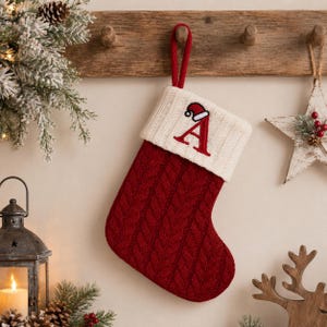 May include: A deep red cable-knit Christmas stocking featuring a cream-colored ribbed cuff. The cuff is embroidered with a large red letter A topped with a small Santa hat. The stocking includes a matching red fabric loop for hanging purposes.