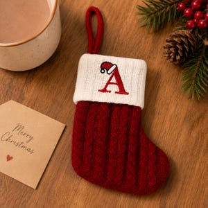May include: A small, deep red cable-knit Christmas stocking with a white ribbed cuff. The cuff features an embroidered red letter A topped with a miniature Santa hat. A red fabric loop is attached to the top for hanging this holiday decoration.