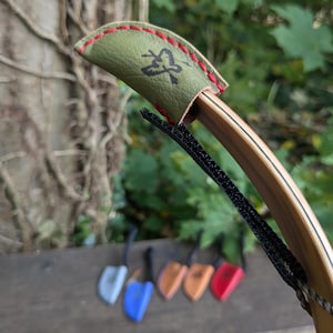 May include: An olive green leather bow tip protector with red stitching and a black arrow logo, attached to the end of a wooden recurve bow with a black strap. In the background, several other bow tip protectors in various colors are visible.