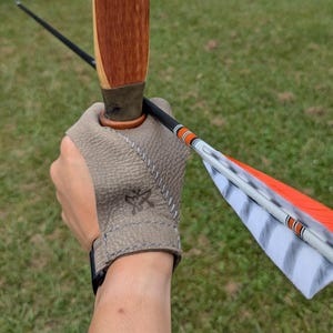 May include: A textured beige leather archery hand guard featuring a small embossed logo and white stitching. The guard is positioned on a hand holding a wooden bow and a black arrow with orange and white striped fletching and a white nock.