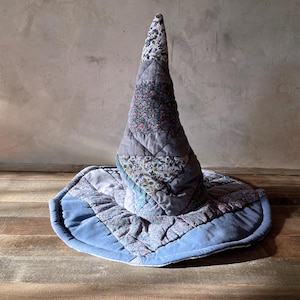 May include: A conical hat constructed from quilted fabric patches in various shades of blue and grey. The design features a mix of solid blue panels and floral patterned sections, with a wide, circular brim and a tall, pointed crown structure.