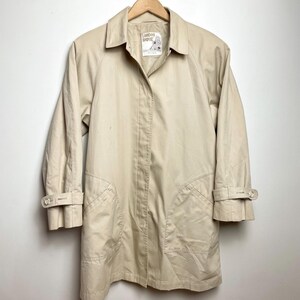 May include: A beige trench coat featuring a pointed collar, long sleeves with buttoned cuff straps, and two large front pockets. The interior label displays the text London Towne. The coat has a button-front placket and a structured, classic silhouette.