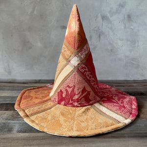 May include: A conical hat constructed from quilted fabric panels in shades of red, orange, and gold. The textile features a woven leaf pattern throughout, with visible stitching lines connecting the various colored sections of the hat's brim and crown.