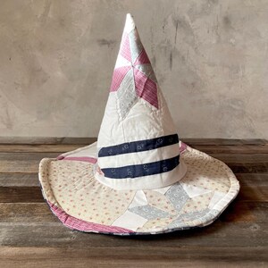 May include: A conical hat constructed from quilted fabric patches in white, red, and navy blue. The design features star-shaped patterns, small floral prints, and gingham checks, with two horizontal navy blue bands encircling the base of the crown.