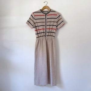 May include: A beige midi dress featuring short sleeves and a round neckline. The bodice has horizontal stripes in red, black, and brown, with a central vertical button placket. The waist is elasticized, leading to a solid beige skirt section.