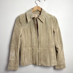 May include: A beige suede jacket featuring a full-length front zipper, a pointed collar, and long sleeves. The garment includes vertical paneling details, a horizontal waist seam, and two side pockets, all finished with tonal stitching throughout.