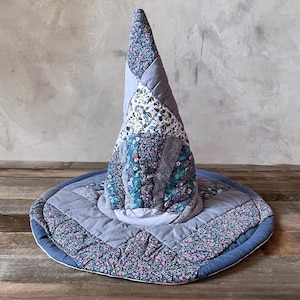 May include: A conical hat constructed from quilted fabric patches in various shades of blue, grey, and floral patterns. The design features a wide, circular brim and a tall, pointed crown, all assembled with visible stitching throughout the textile.