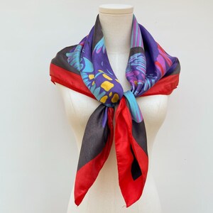 May include: A square scarf featuring a vibrant pattern of purple, blue, and yellow abstract shapes against a dark background, finished with a bold red border. The scarf is tied in a knot at the center, displaying a smooth, satin-like texture.