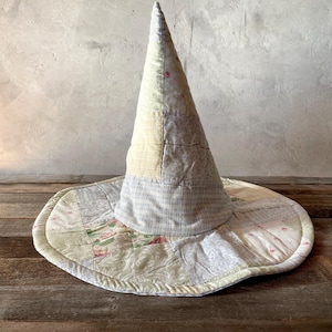May include: A conical hat constructed from various fabric patches in muted shades of cream, beige, light gray, and soft pink. The patchwork design features a mix of floral, plaid, and solid textures, forming a wide, circular brim and a tall crown.
