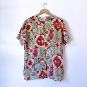 May include: A short-sleeved button-down shirt featuring a patchwork design with red, green, and cream-colored geometric and floral patterns. The shirt has a mandarin collar, a front placket with four toggle-style closures, and side slits at the hem.