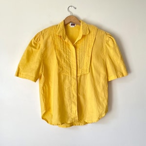 May include: A bright yellow short-sleeved button-down shirt featuring a classic collar and vertical pleat detailing on the front chest panel. The garment has a straight hemline and is displayed on a wooden hanger against a plain background.