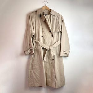 May include: A beige trench coat with a double-breasted design, a belt, and buttoned cuffs. The coat is hanging on a wooden hanger against a white background. The coat's length extends to the mid-calf.