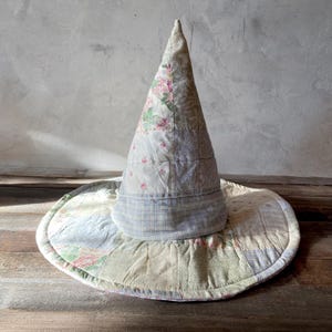 May include: A conical hat constructed from various quilted fabric patches in muted tones of cream, sage green, soft blue, and dusty pink. The patchwork features floral patterns, subtle plaids, and solid textures, forming a wide, circular brim.