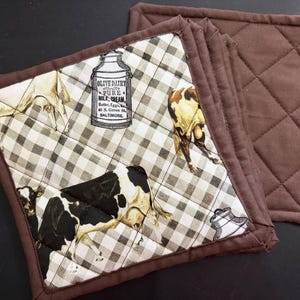 May include: A set of square quilted pot holders featuring a brown and white buffalo check pattern with illustrations of cows and vintage milk cans. The text on the milk can reads Olive Dairy Pure Milk Cream Butter Eggs 46 S. Green St. Baltimore.