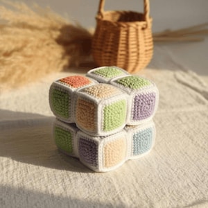 Op de afbeelding: Een zacht, gehaakt kubusspeeltje met een raster van kleine, vierkante panelen in pasteltinten zoals perzik, lichtgroen, lavendel en babyblauw. Elk afzonderlijk vierkant is omlijnd met wit garen, wat zorgt voor een gestructureerd, veelkleurig blokontwerp.