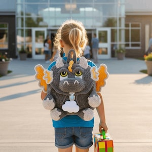 May include: A dark gray crocheted dragon backpack featuring large yellow and orange wings, light gray horns, and a white cloud-shaped tag attached to the silver zipper pull. The dragon face has prominent green eyes and a textured yarn construction.