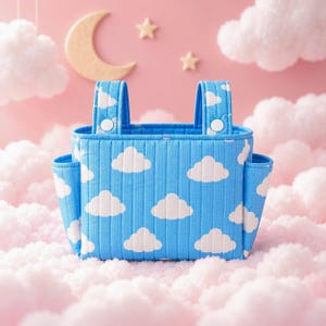 May include: A quilted blue fabric storage caddy featuring a white cloud pattern. The rectangular organizer includes two vertical straps with white snap buttons at the top and two side pockets, providing a structured container for small items.
