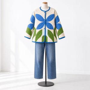 May include: A quilted jacket featuring a cream base with a large blue floral pattern and green leaf motifs, finished with blue trim and buttons. The jacket is paired with straight-leg blue denim jeans on a display mannequin.
