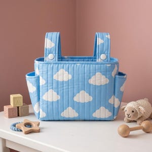 May include: A light blue quilted storage basket featuring a white cloud pattern, two vertical handles with white snap buttons, and two side pockets. The basket is rectangular with vertical stitching and a structured, upright design.