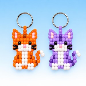 May include: Two cat-shaped keychains crafted from small square beads. One keychain is orange with white accents, and the other is purple with white accents. Both feature black bead eyes, pink bead noses, and silver-toned metal key rings at the top.