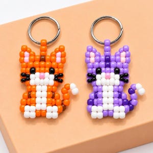 Op de afbeelding: Twee katvormige sleutelhangers gemaakt van kleine plastic kralen. De ene is oranje met witte accenten, de andere is paars met witte accenten. Beide hebben zwarte kraaloogjes, een roze kraalneus en zilverkleurige metalen sleutelringen.