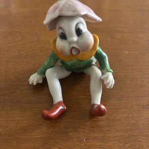 Può includere: Una stravagante statuetta in ceramica di un personaggio a forma di fungo. Presenta un viso bianco con grandi occhi, una testa a forma di fiore rosa e bianco e un abito verde. La statuetta ha gambe bianche, scarpe rosse e un colletto giallo.
