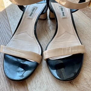 May include: A pair of Miu Miu sandals featuring a glossy beige patent leather toe strap and ankle strap with a gold-tone buckle. The footbed is light beige leather with the Miu Miu logo, paired with a black patent leather heel and trim.