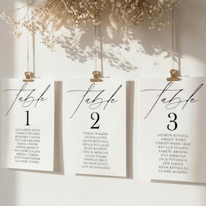 May include: Three white rectangular seating chart cards hanging from gold metal clips. Each card features the word Table in black script, a large number, and a list of names in black serif font. The cards are suspended by thin strings.
