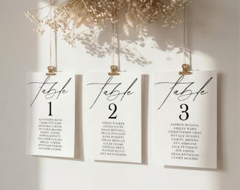 May include: Three white rectangular seating chart cards hanging from gold metal clips. Each card features the word Table in black script, a large number, and a list of names in black serif font. The cards are suspended by thin strings.