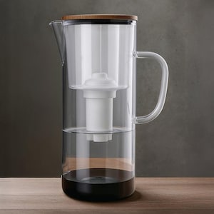 Puede incluir: Una jarra filtrante de agua de vidrio transparente con base negra y tapa de madera. La jarra tiene un filtro blanco en el interior y un asa curva. El agua en el interior es clara. La jarra está sobre una superficie de madera.