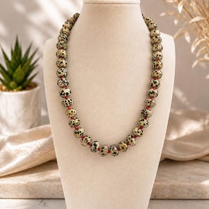 Puede incluir: Un collar de cuentas compuesto por cuentas redondas de jaspe dálmata color crema con manchas negras, intercaladas con pequeñas cuentas espaciadoras de color rojo vibrante. El collar se exhibe sobre un busto de tono neutro en una disposición circular uniforme.