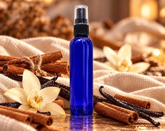 Pode incluir: Um frasco pulverizador cilíndrico de vidro azul cobalto com uma tampa de bomba atomizadora de plástico preto, posicionado na vertical entre paus de canela castanhos, vagens de baunilha escuras e uma flor de baunilha creme.