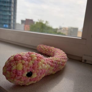 Könnte beinhalten: Ein handgefertigtes Häkelschlange-Spielzeug in Rosa und Gelb mit einem schwarzen Auge. Die Plüschschlange liegt auf einer Fensterbank, mit Blick auf die Stadt im Hintergrund. Ein niedliches und kuscheliges Spielzeug.