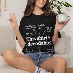 May include: A black t-shirt featuring white text that reads "This shirt is decodable." The design includes various arrows pointing to the text with labels such as "Closed Syllable = short vowel sound," "Digraphs," "r-controlled," and "Latin-based."
