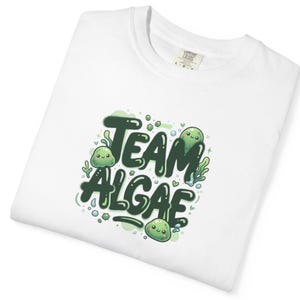 Peut inclure: Un t-shirt blanc avec une impression graphique portant le texte "TEAM ALGAE" en lettres bulles vert foncé. Le motif comprend de petits personnages en forme de goutte verts souriants, des accents d'algues vert clair et des bulles.