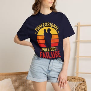 Könnte beinhalten: Ein marineblaues Kurzarm-T-Shirt mit einer Grafik eines Sonnenuntergangs in Gelb, Orange und Rot. Das Design zeigt die Silhouette einer Person, die ein Kind hält, mit dem Text "PROFESSIONAL PULL OUT FAILURE" in gelber und roter Schrift.