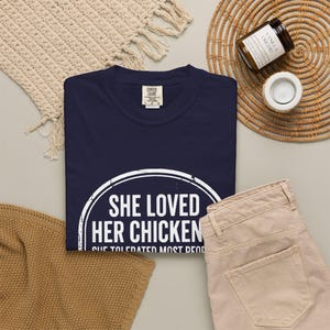 May include: A navy blue short-sleeve t-shirt featuring a white circular graphic with the text "SHE LOVED HER CHICKENS SHE TOLERATED MOST PEOPLE" in a distressed font. The shirt has a Comfort Colors brand label at the inner neckline.