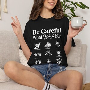 以下が含まれることがあります： Be Careful What You Wish Forという白いテキストと、富、名声、世界平和など様々な願い事を描いた一連の小さなグラフィックイラスト、および追加のテキストラベルがプリントされた黒い半袖Tシャツ。