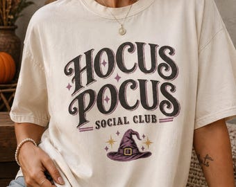Könnte beinhalten: Ein elfenbeinfarbenes Kurzarm-T-Shirt mit dem schwarzen Schriftzug HOCUS POCUS SOCIAL CLUB und einer Grafik eines violetten Hexenhutes mit goldenen Sternakzenten. Das Shirt hat einen Rundhalsausschnitt und eine lockere Passform mit Frontdruck.