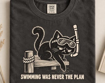 Könnte beinhalten: Ein anthrazitfarbenes T-Shirt mit einer Grafik einer schwarzen Katze, die Schnorchelausrüstung trägt und auf einem Holzsteg sitzt. Die Katze berührt das Wasser mit einer Pfote. Der Text darunter lautet: "SWIMMING WAS NEVER THE PLAN". Comfort Colors Etikett.