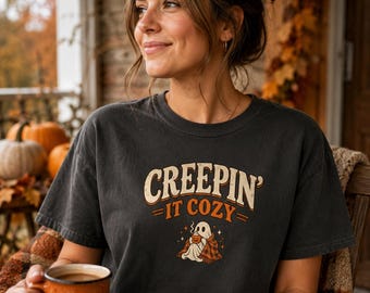 Könnte beinhalten: Ein anthrazitfarbenes Kurzarm-T-Shirt mit dem Schriftzug "CREEPIN' IT COZY" in cremefarbenen und orangefarbenen Buchstaben. Unter dem Text befindet sich eine Grafik eines Geistes, der in eine karierte Decke gehüllt ist und eine dampfende Kürbistasse hält.