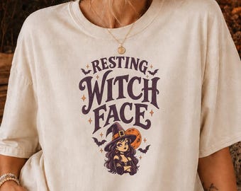 Könnte beinhalten: Ein cremefarbenes Kurzarm-T-Shirt mit dem violetten Schriftzug Resting Witch Face über einer Grafik einer Zeichentrickhexe mit Hut, umgeben von kleinen Fledermäusen und einem großen orangefarbenen Mond, akzentuiert durch Sterne auf der Vorderseite.