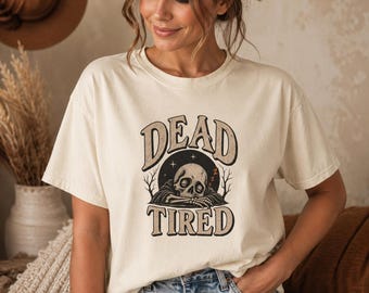 Könnte beinhalten: Ein cremefarbenes Kurzarm-T-Shirt mit einem Grafikdruck eines schlafenden Schädels, der seinen Kopf auf die Arme stützt. Der Text "DEAD TIRED" ist in großen, fetten, bogenförmigen Buchstaben über und unter der Illustration gedruckt.