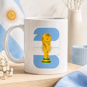 K&ouml;nnte beinhalten: Wei&szlig;e Keramiktasse mit einem stilisierten Design der argentinischen Flagge und einer goldenen Troph&auml;e. Die Tasse hat einen Henkel und steht auf einer Holzoberfl&auml;che, mit einer Flagge im Hintergrund. Die Troph&auml;e hat einen gr&uuml;nen und gelben Sockel.