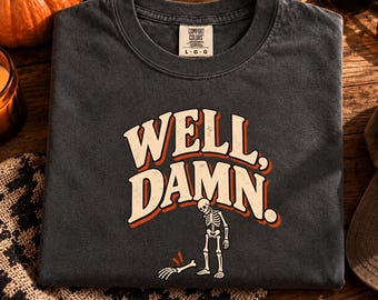 Könnte beinhalten: Ein anthrazitfarbenes T-Shirt mit dem Schriftzug WELL, DAMN. in cremefarbener Schrift mit orangefarbenen und schwarzen Umrandungen. Unter dem Text befindet sich eine Grafik eines Skeletts, das neben seiner abgetrennten Hand steht.